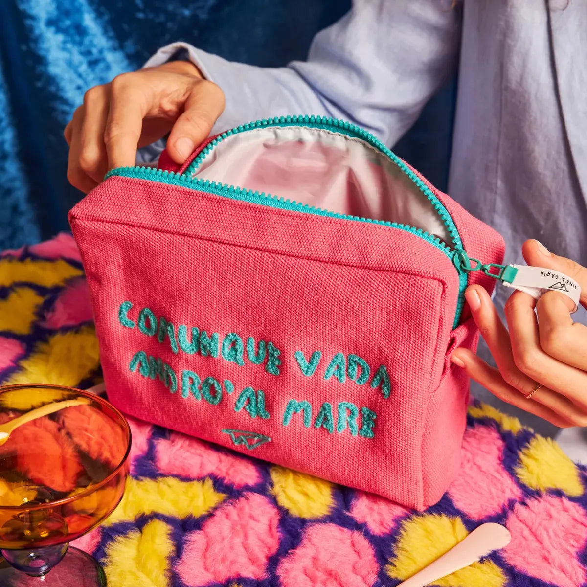 Mano che apre la zip azzurra della trousse fucsia con la scritta azzurra "Comunque vada andrò al mare"