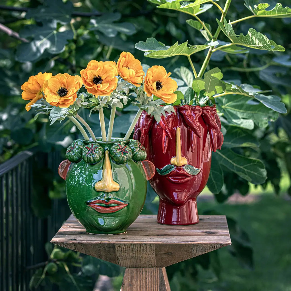 Due vasi con volto maschile raffigurato appoggiati su un tavolo di legno. Entrambi i vasi hanno una corona di verdura verde e rossa. Fiori e piante inseriti nei vasi.