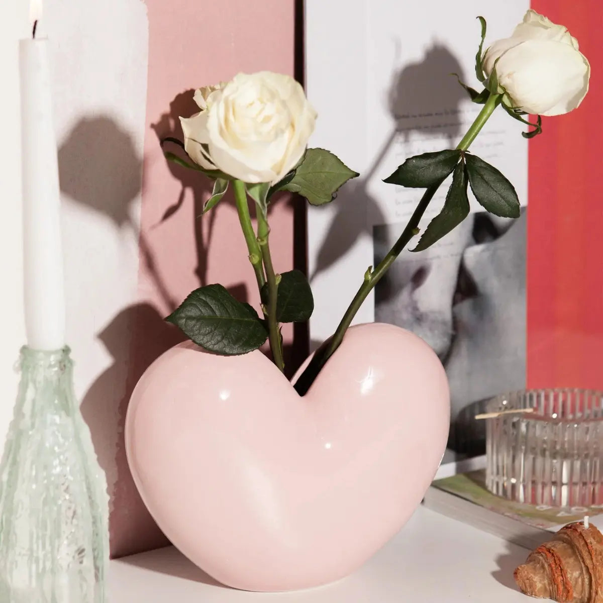 Vaso a forma di cuore rosa di DOIY con all'interno due rose bianche. Il vaso è appoggiato su una mensola con altri oggetti intorno