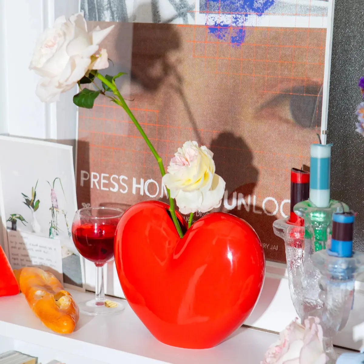 Vaso a forma di cuore rosso di DOIY con all'interno due rose bianche. Il vaso è appoggiato su una mensola con altri oggetti intorno