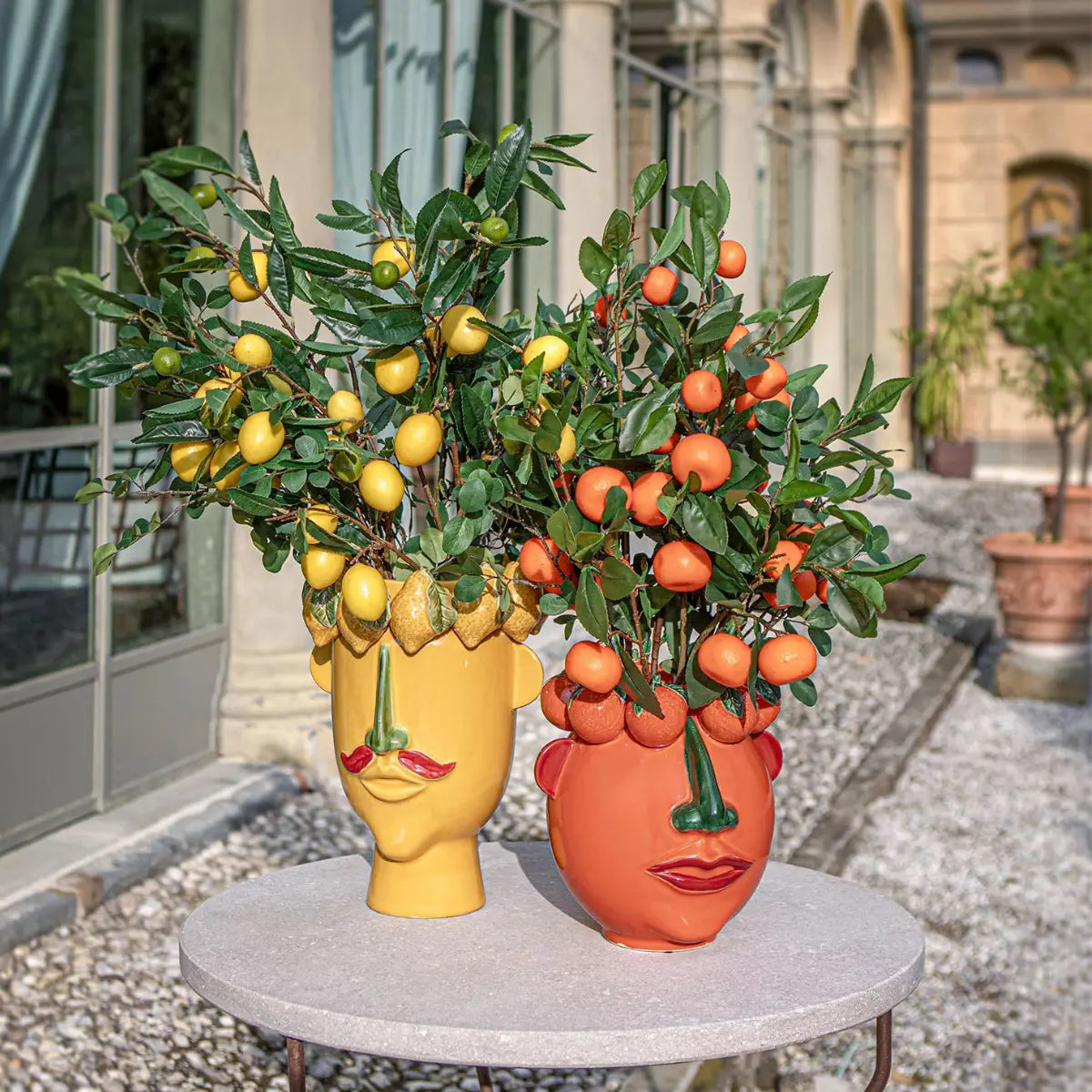Due vasi con volti maschili e corona di frutta posizionati su un tavolo in cemento con rami di limoni e mandarini inseriti.