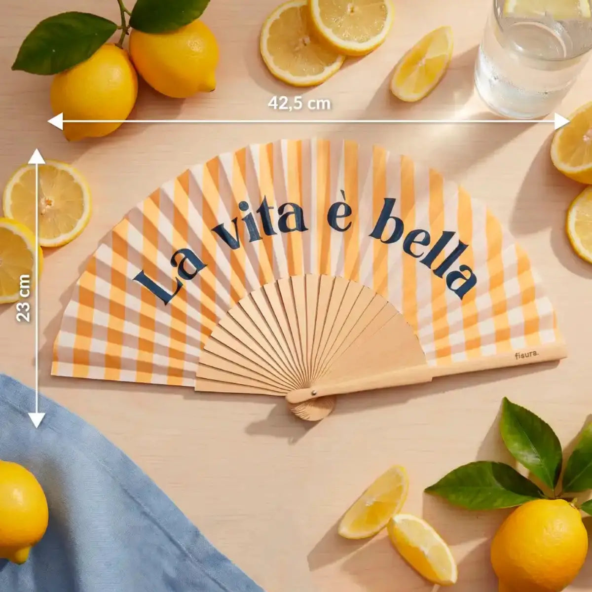 Ventaglio con base a righe gialle e bianche e scritta blu "La vita è bella". Ventaglio appoggiato su un tavolo con misure segnate in bianco con frecce
