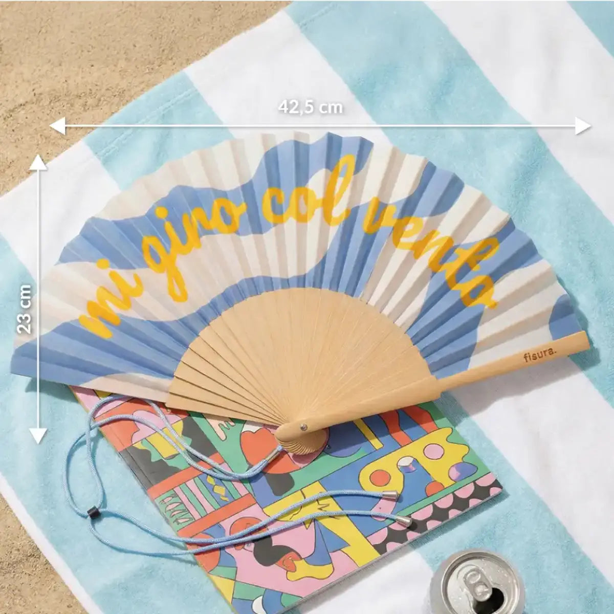 Ventaglio con sfondo fantasia azzurra e bianca con scritta gialla "Mi giro col vento" appoggiato su un telo mare in spiaggia. Le misure sono segnate con una freccia e un numero in bianco 