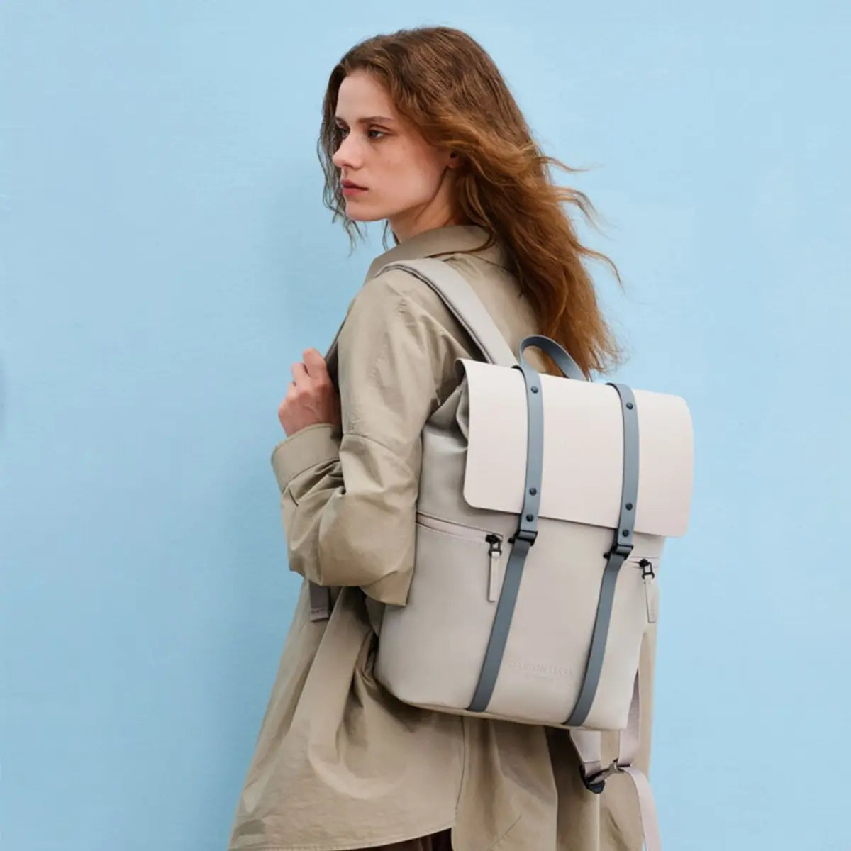 Ragazza che indossa lo zaino con patta nella colorazione tortora. Sfondo azzurro