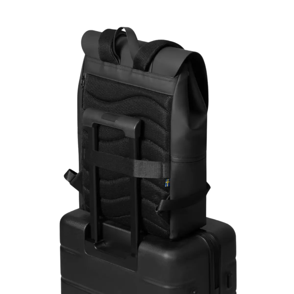 Zaino nero con patta posizionato sopra una valigia nera e fissato con l'apposito gancio. Sfondo bianco