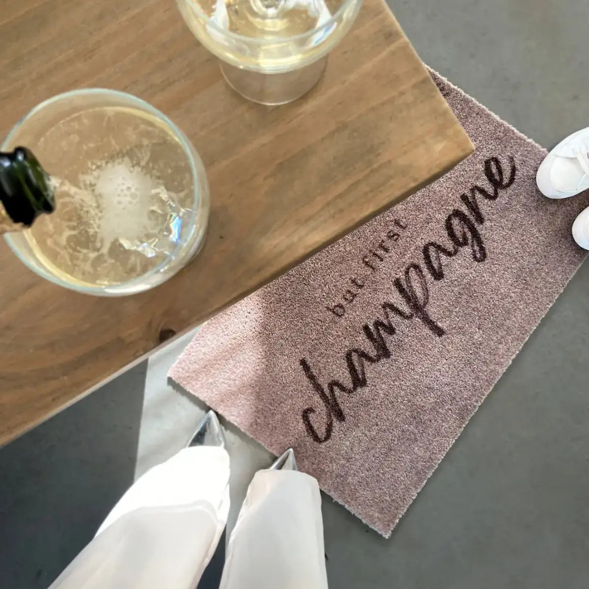 Zerbino beige con scritta but first champagne su un pavimento grigio con due calici di champagne sul tavolo