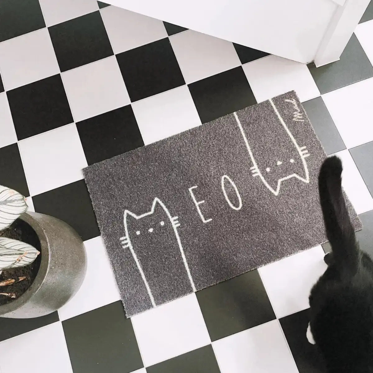 Zerbino grigio con gatti stilizzati su un pavimento di piastrelle e un gatto nero 